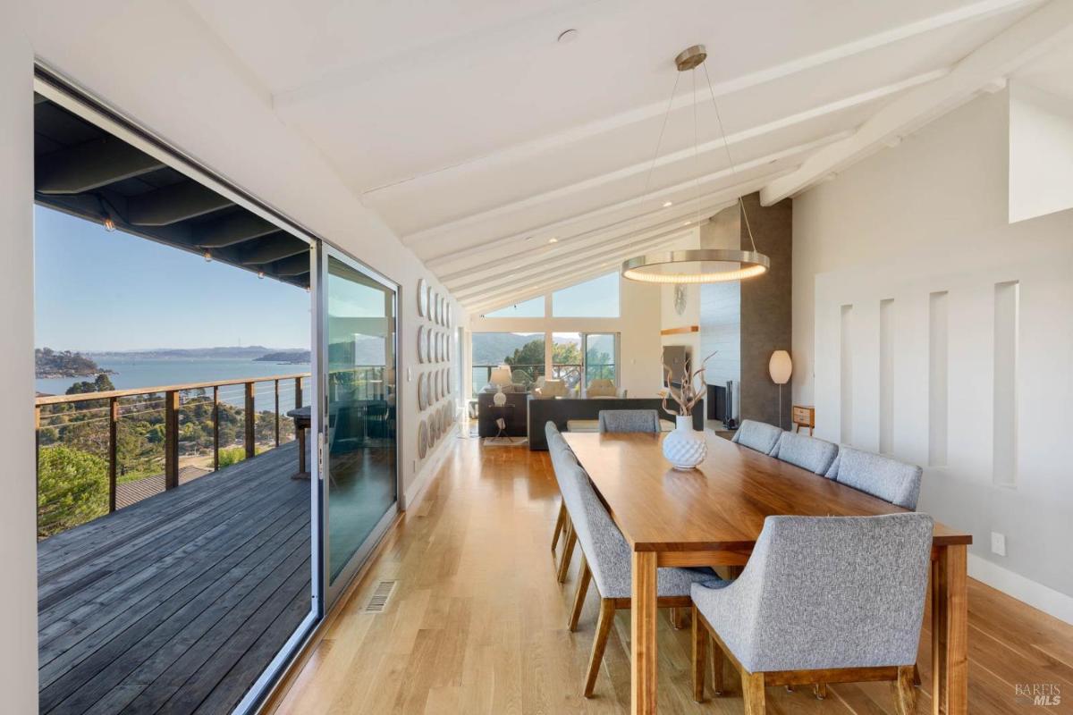 A dining area features a long wooden table with gray chairs and large windows overlooking a bay.