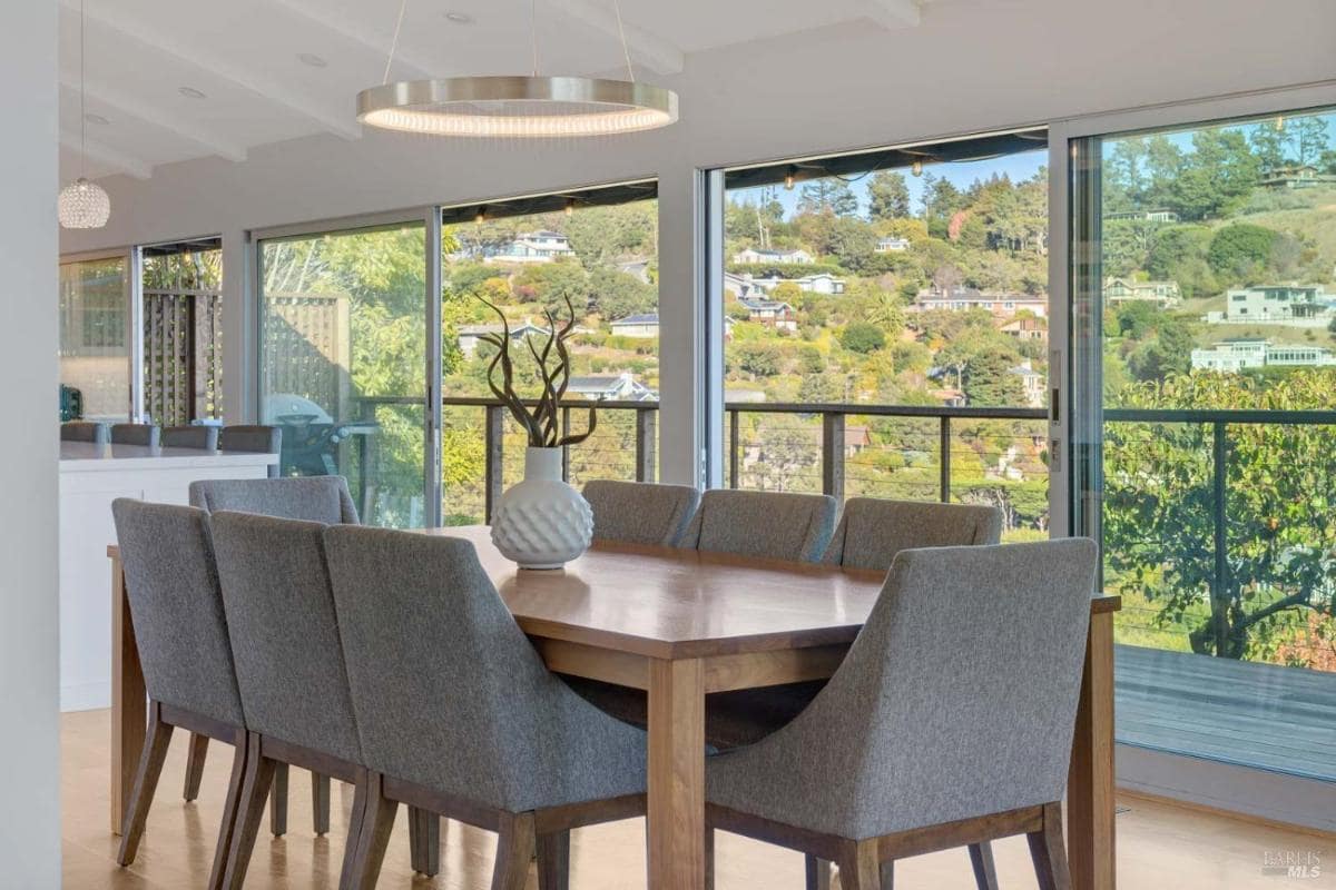 A dining table with gray chairs is positioned near large windows that view a hillside with houses.