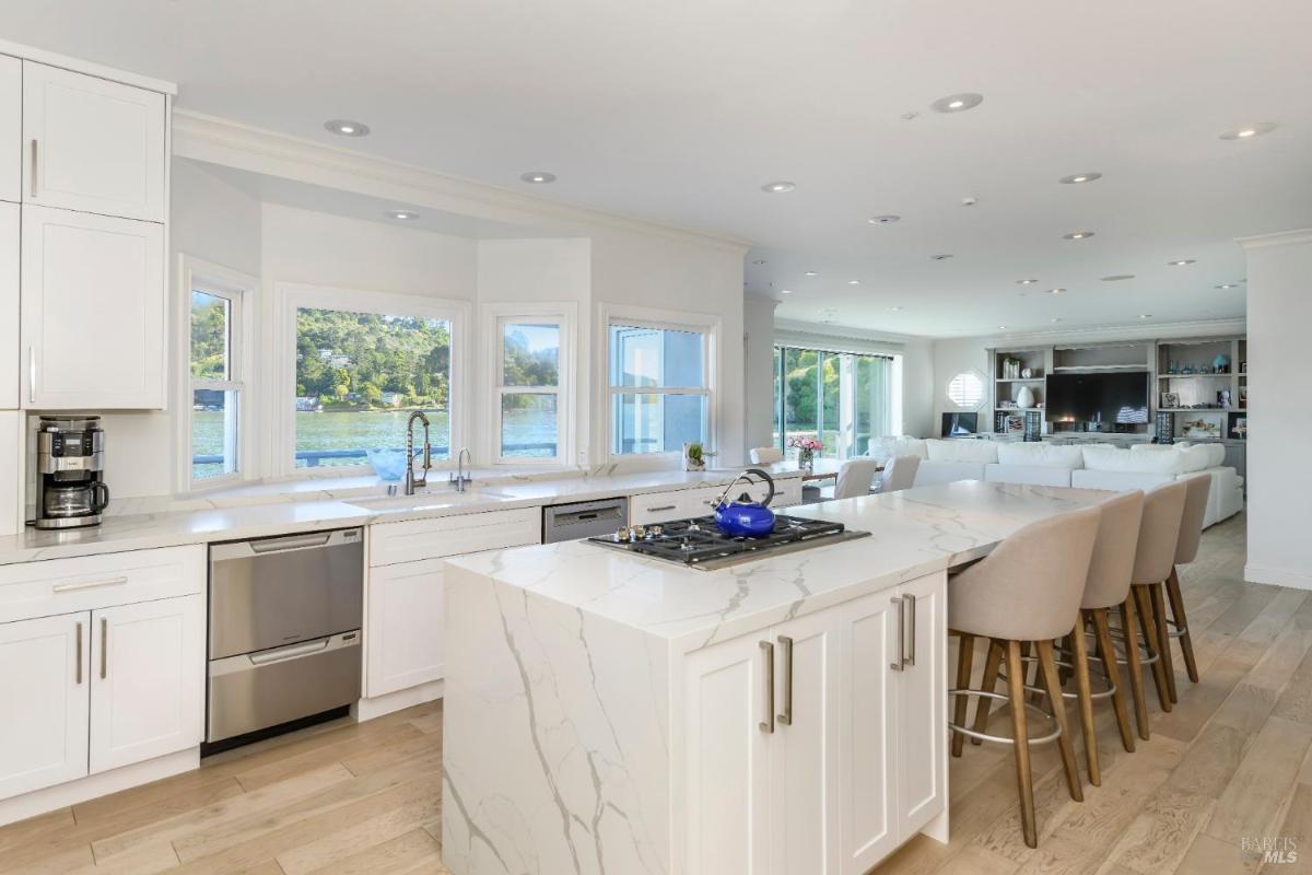 A modern kitchen with a white island, barstool seating, and large windows overlooking the water.
