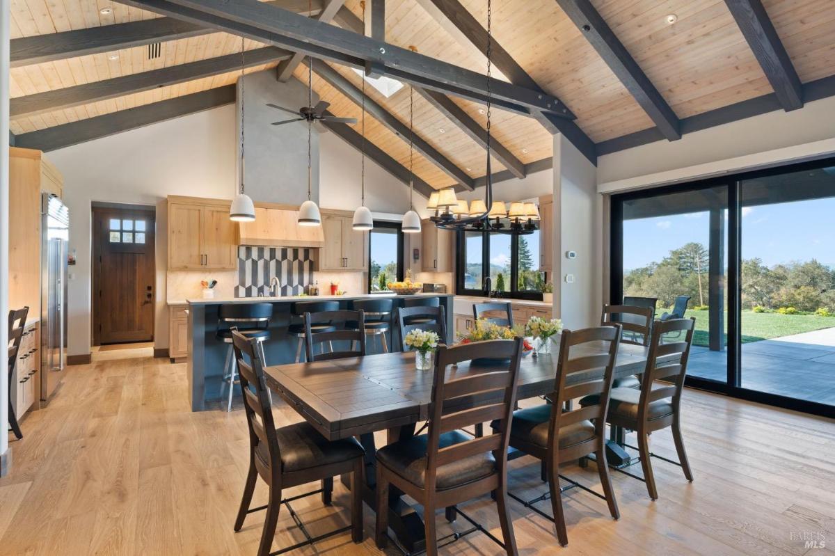 View of a wooden dining table, a chandelier, and the kitchen in the background with pendant lights, an island with barstools, and wooden cabinetry.