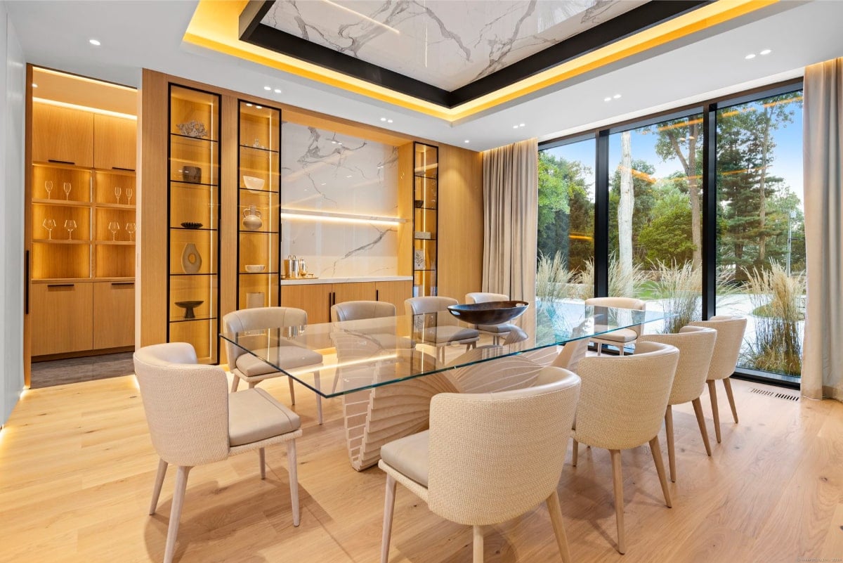 The room features a glass dining table with white chairs, built-in cabinetry, and large windows offering a view of greenery. 