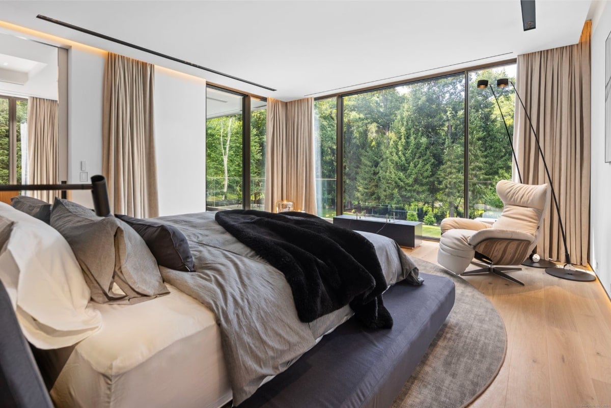Bedroom with floor-to-ceiling windows offering a view of a forest or wooded area. The room is furnished with a large bed, a comfortable armchair, and floor-length curtains.