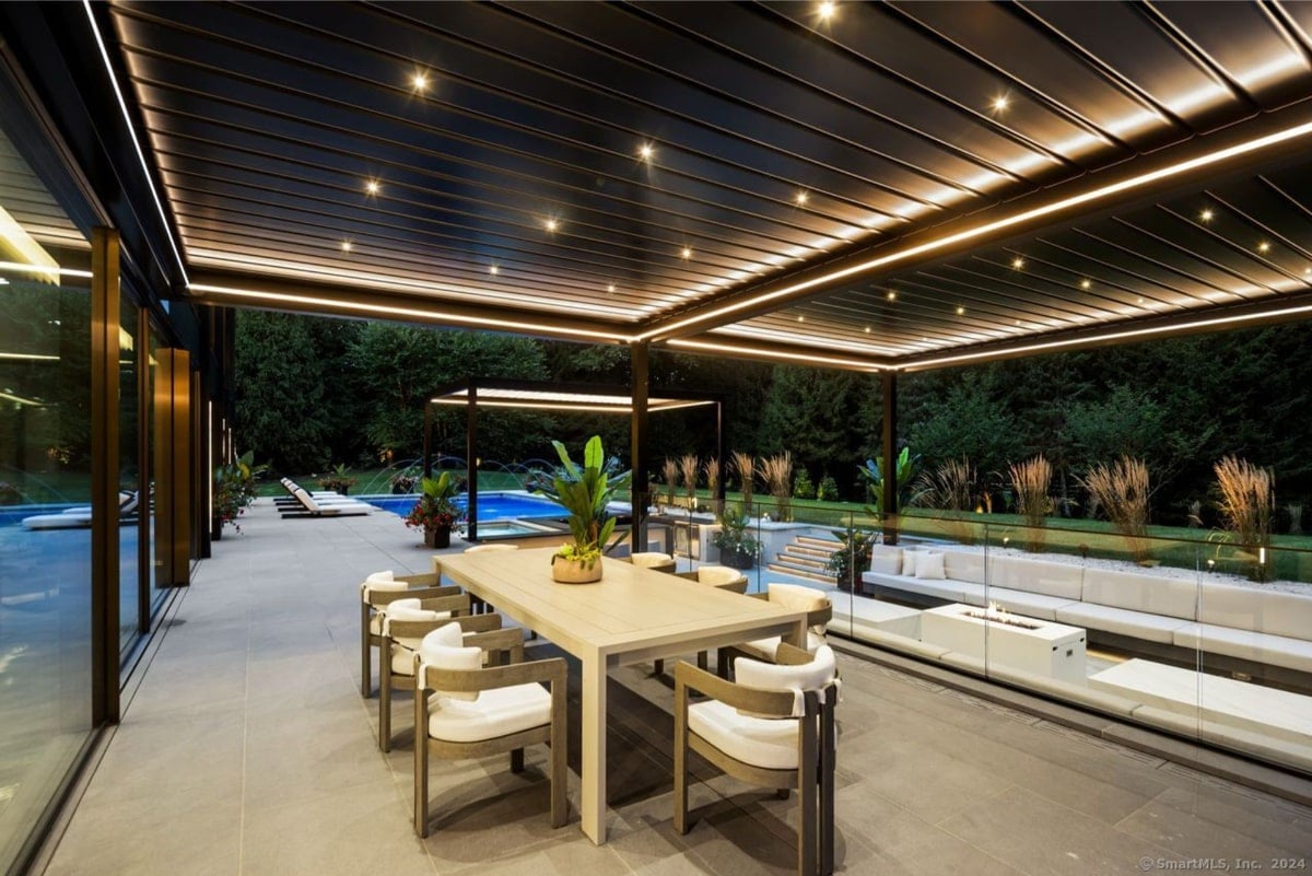 An outdoor dining area at night, situated next to a swimming pool. It features a long, light-colored table with matching chairs under a pergola with integrated lighting. 