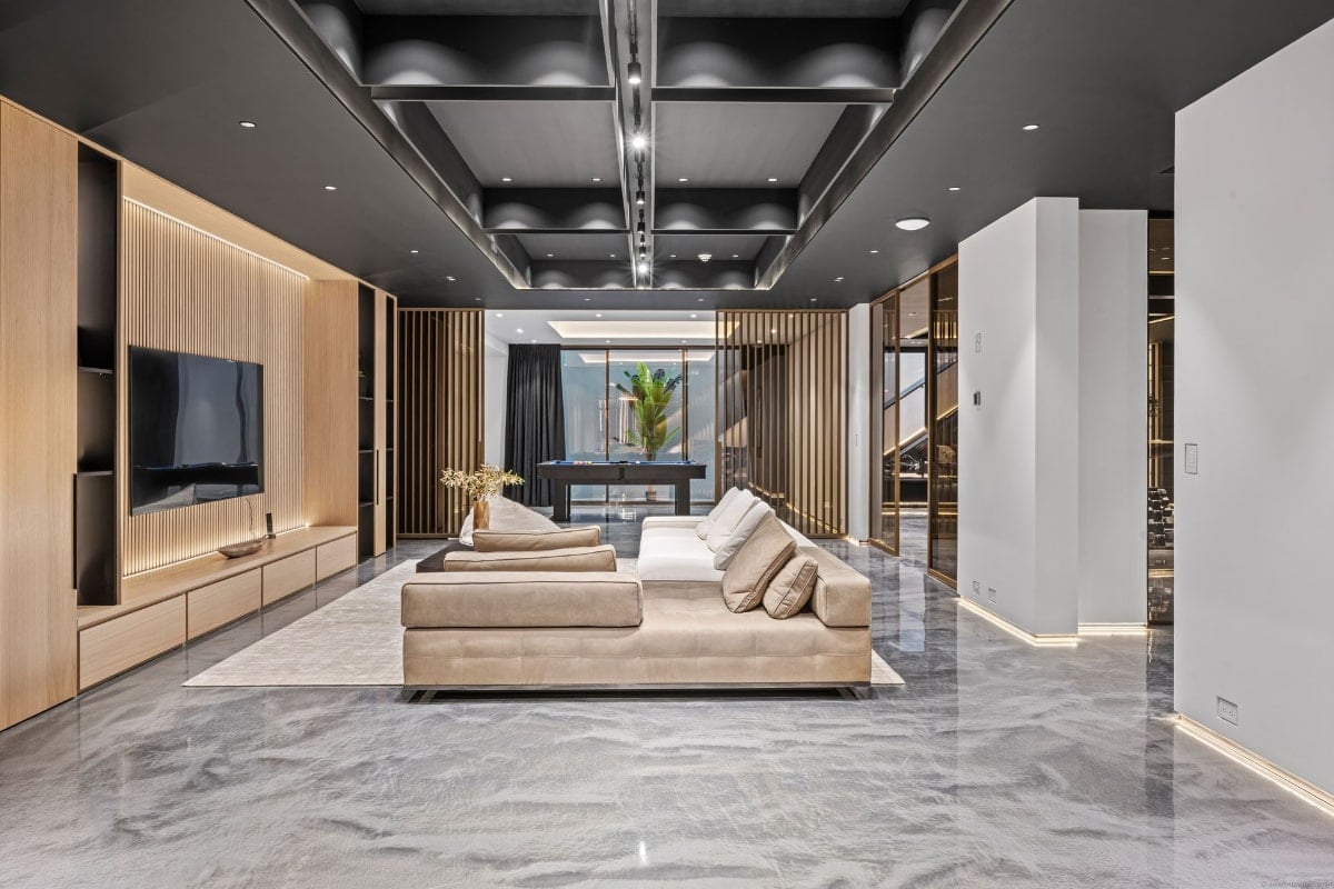 Living area with a low-slung white sofa as the central feature. The room has a dark ceiling, light-colored walls, and a polished floor. A glimpse of a game room or similar space is visible in the background.