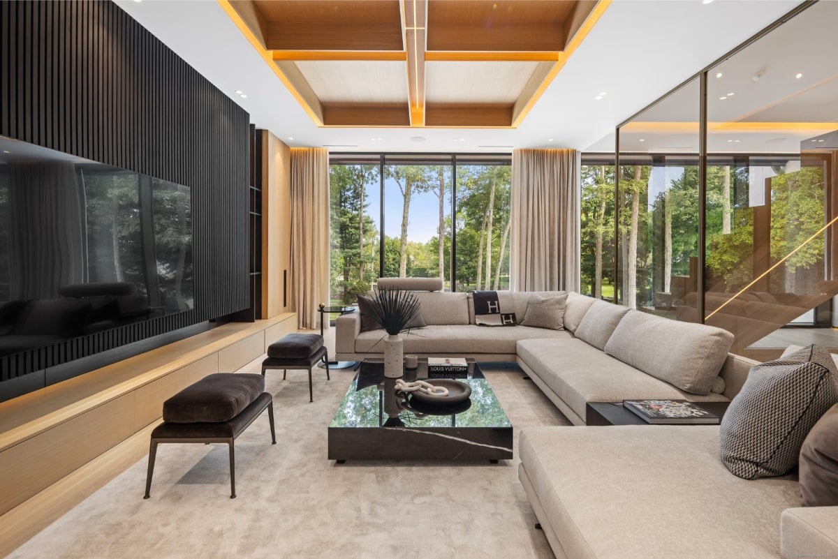 A living room with a large sectional sofa, dark wood accents, and floor-to-ceiling windows overlooking trees.