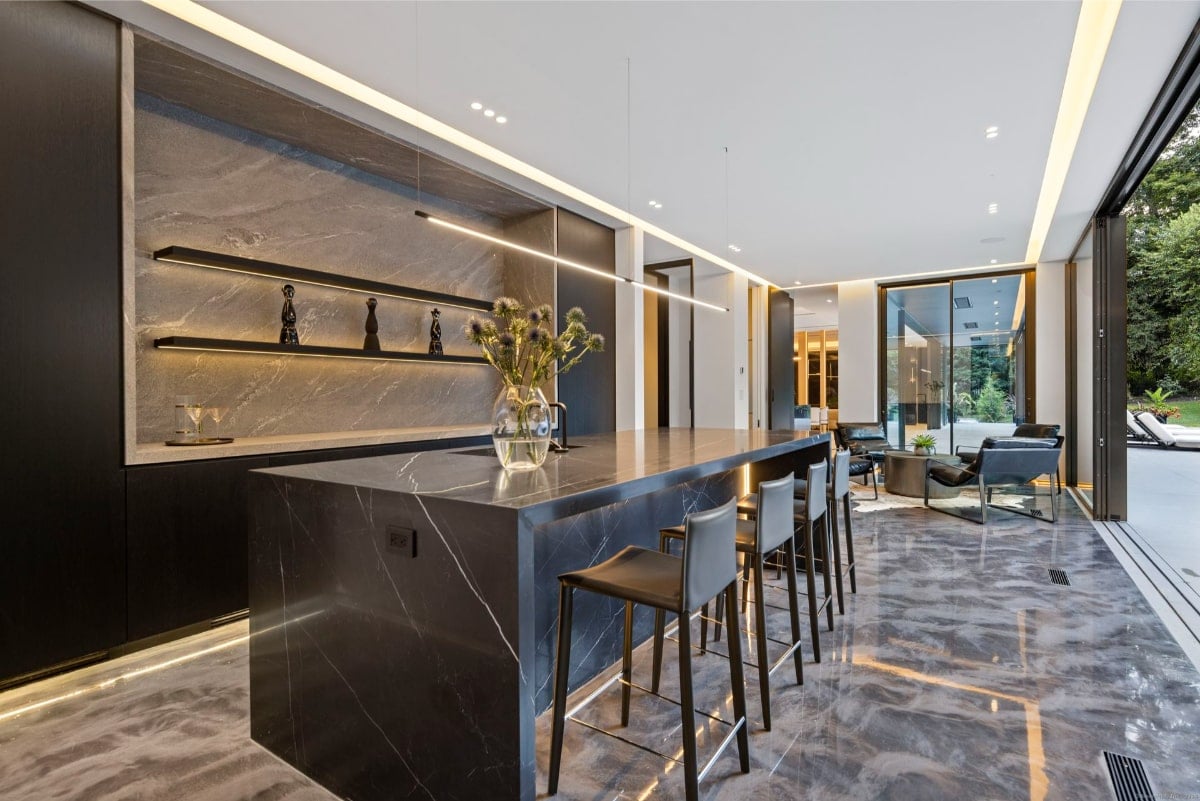 Bar area with a large island featuring a dark marble countertop. The island has a distinctive blue edge detail. There are several bar stools around the island. The backsplash is also marble, and there's a minimalist shelf with decorative objects. The floor is a polished, light gray material.