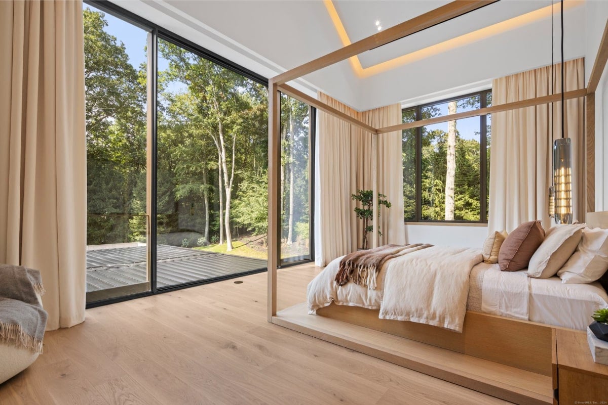 A bedroom with a four-poster bed and floor-to-ceiling windows that offer a view of trees and what appears to be a deck or patio. The room is decorated in a minimalist style with light-colored walls and neutral bedding.