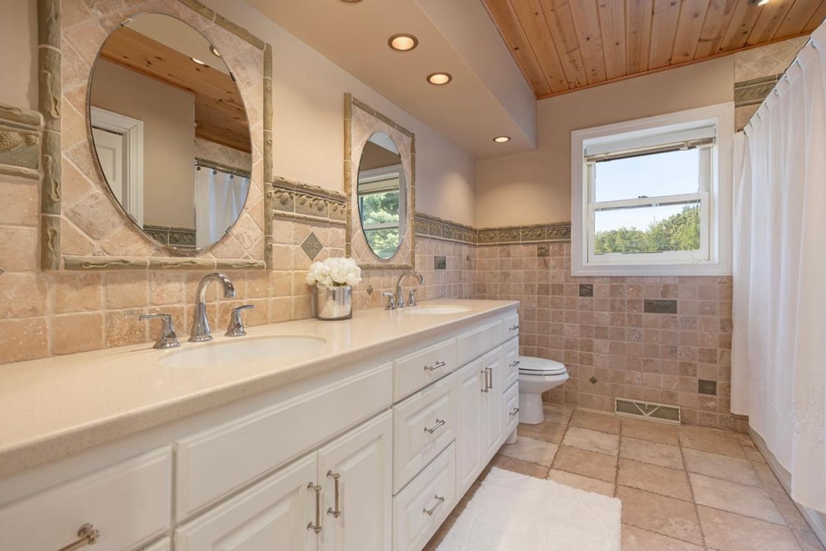 White vanities, oval mirrors, and neutral tile flooring create a bright bathroom.