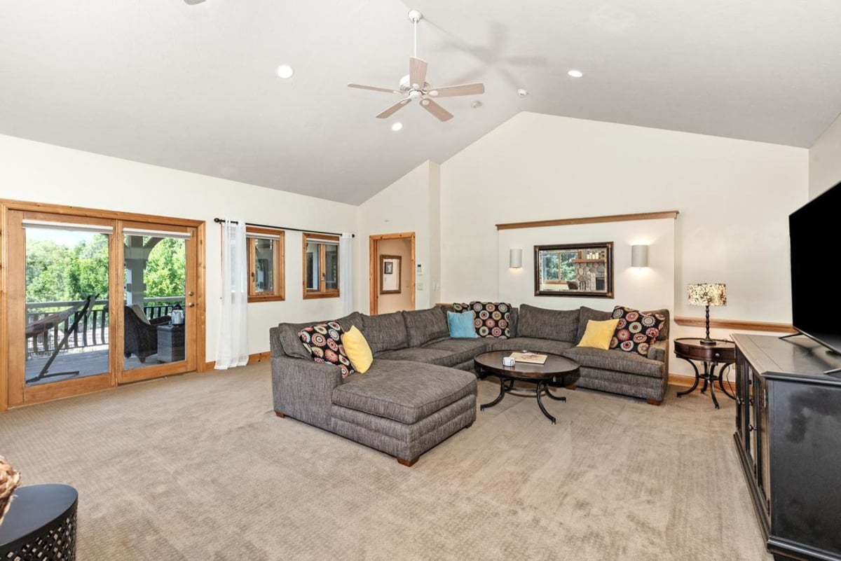 Sectional sofa dominates this spacious room, complemented by high ceilings, a ceiling fan, and access to an outdoor deck.