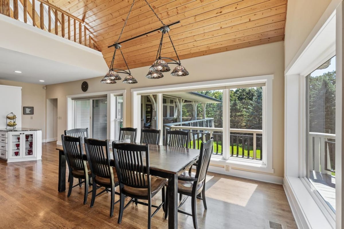 Wood-paneled ceiling, large windows offering scenic views, and a rustic dining table create a warm and inviting atmosphere in this dining area.