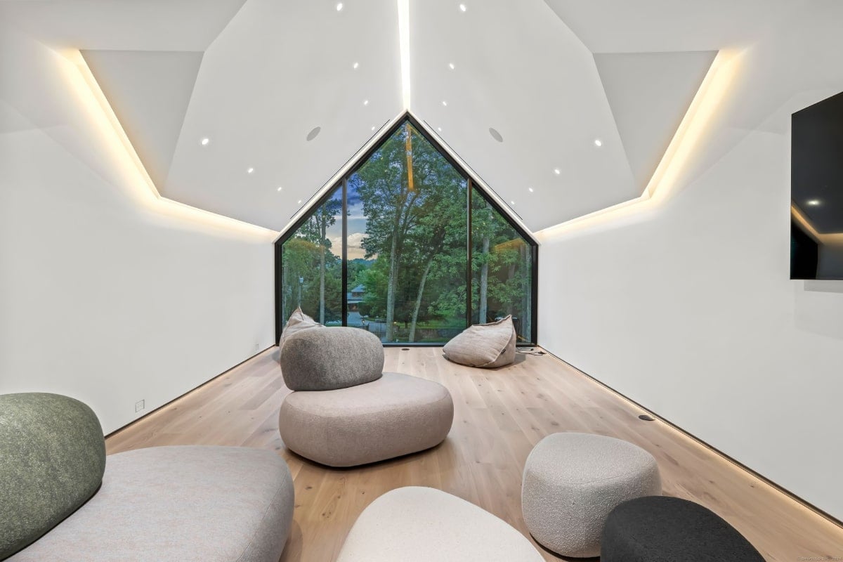 A room with a sloped ceiling and large window offering a view of trees. The room is sparsely furnished with several large, light-colored, organic-shaped seating elements.