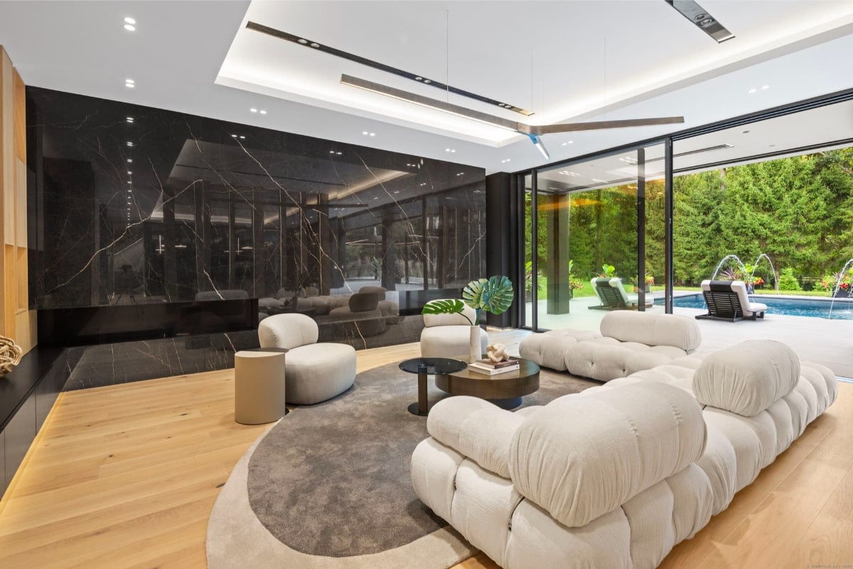 Living room with puffy white sectional sofa. A dark marble wall is a prominent feature, and large windows offer a view of an outdoor pool area with water features.
