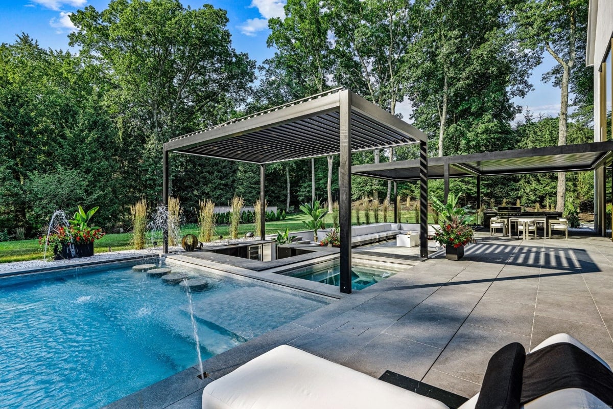Outdoor pool area with multiple levels and water features. There are two large, dark-colored pergolas providing shade over parts of the pool and a patio area with seating. The landscaping includes tall grasses and potted plants