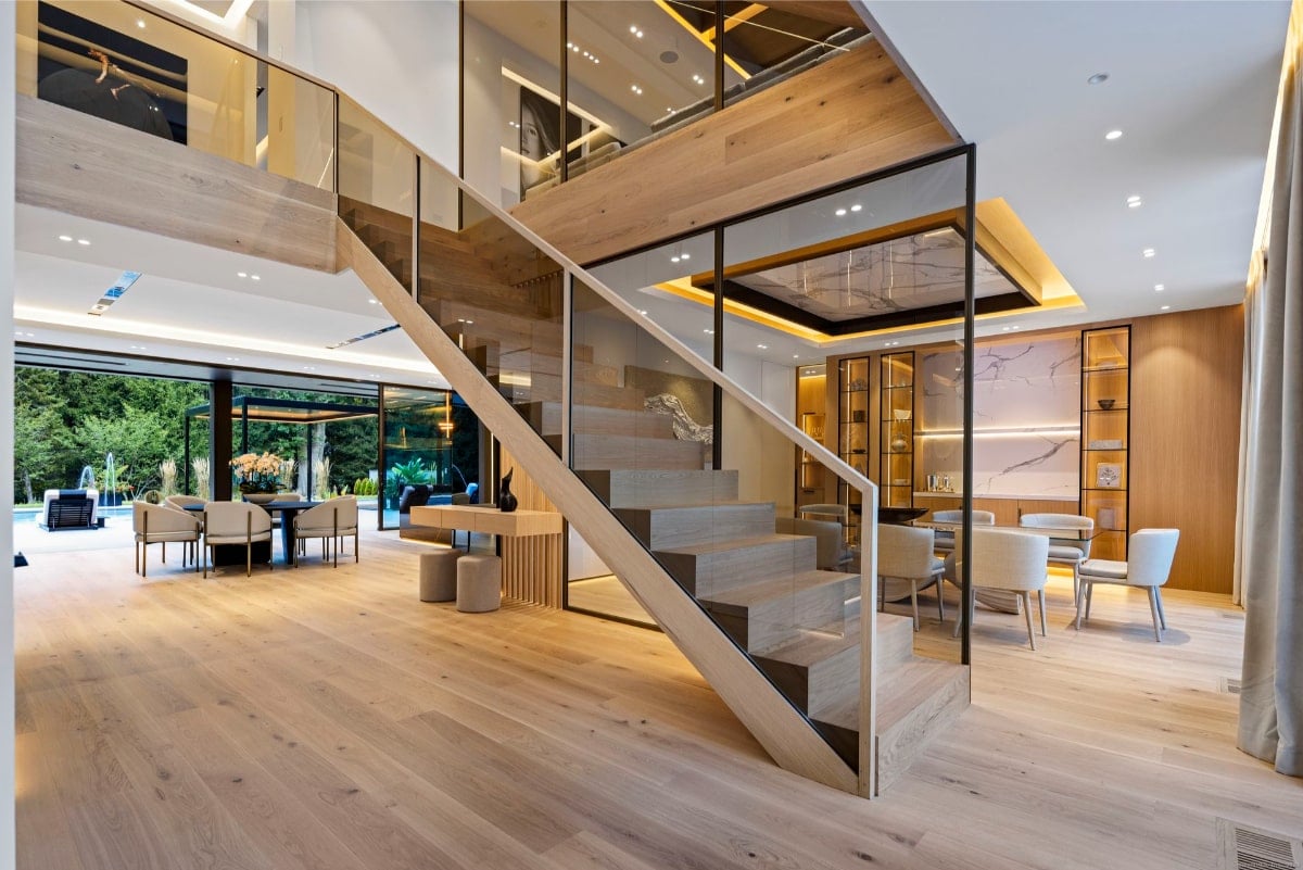 Staircase with glass railings leading to a second floor. The staircase is situated in an open-plan area with light wood floors, and views of a dining area and outdoor space are visible.