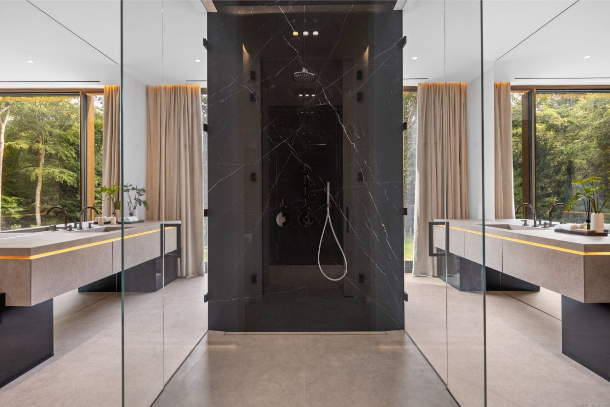  Bathroom with dark-colored shower enclosure as a central feature. The shower is framed by glass, and the walls are marble. There are double vanities on either side of the shower, and large windows offer a view of trees outside.