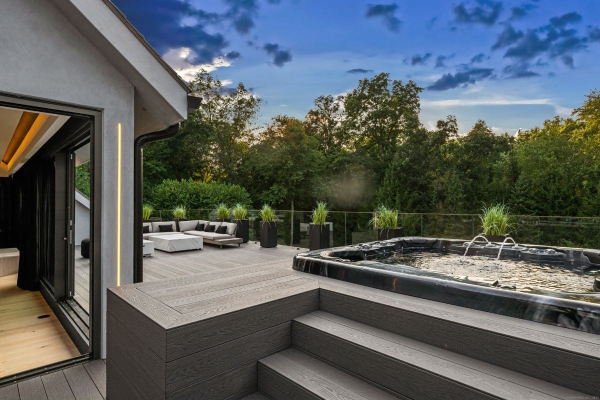 Outdoor space featuring a large hot tub situated on a tiered deck overlooking a wooded area. The deck also has comfortable seating, suggesting a relaxing and upscale environment.