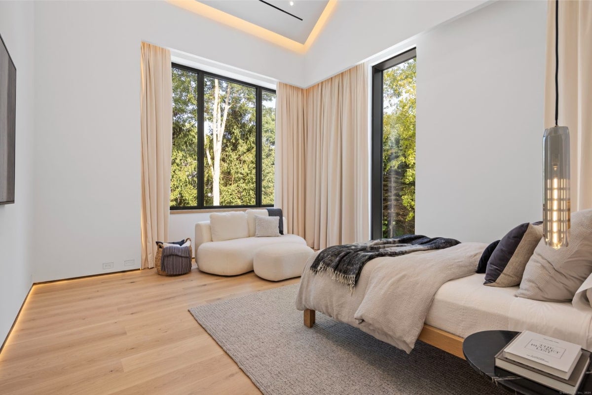 Bedroom with light bedding, a seating area with a white sofa, and floor-to-ceiling windows that provide ample natural light and views of trees. The floor is light-colored wood.
