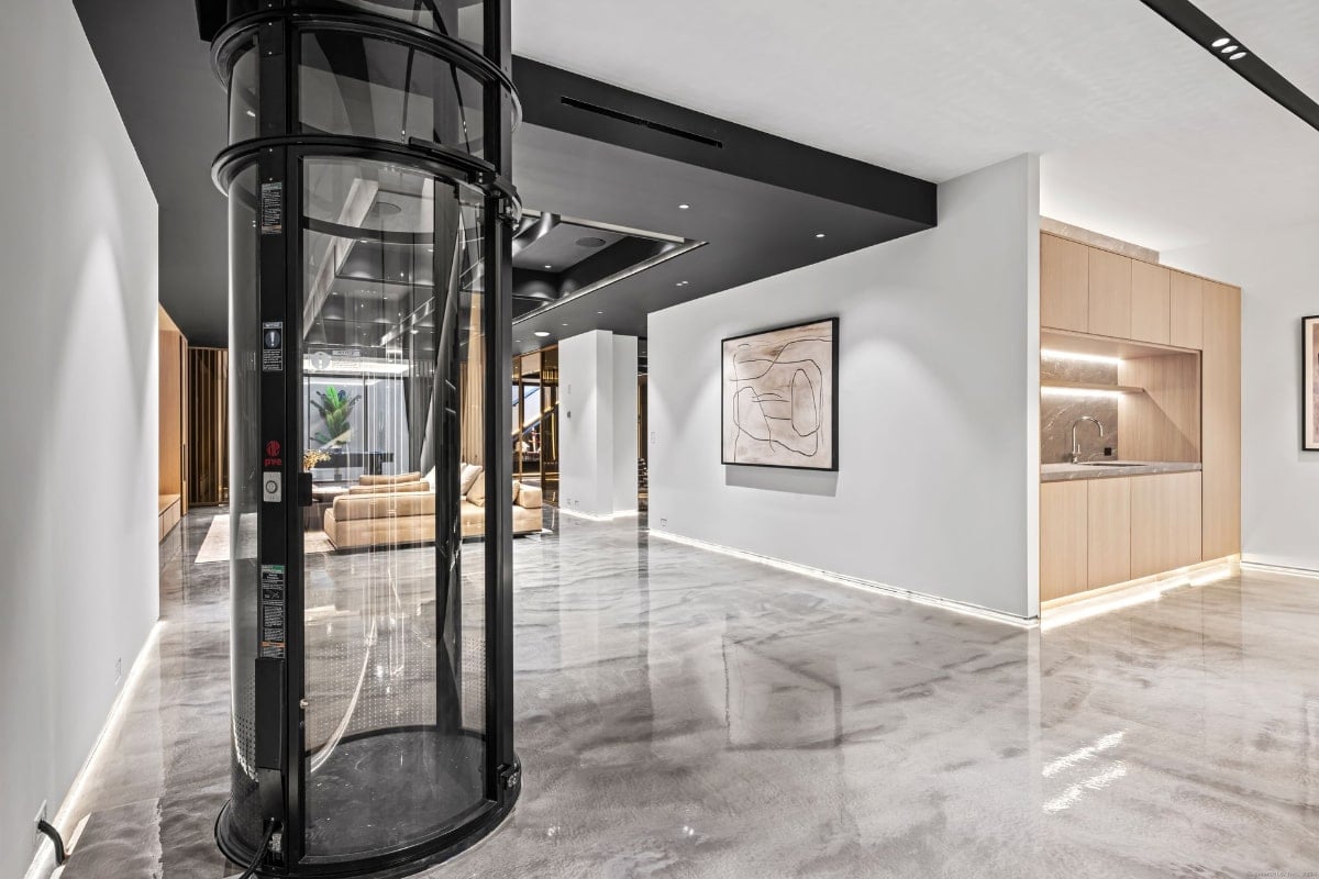 Interior space with a unique circular glass elevator as a central feature. There's a minimalist kitchenette and artwork on the walls. The floor appears to be polished concrete.