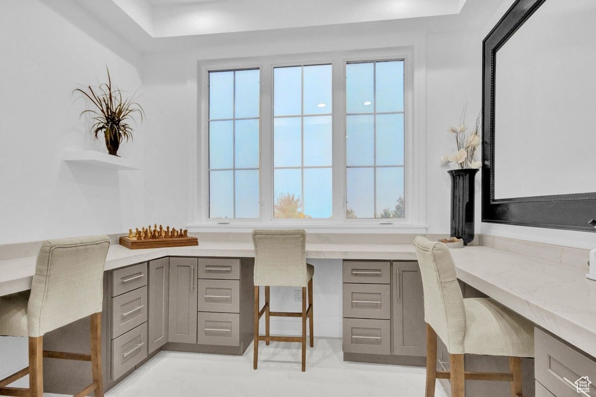 A spacious home office is presented in this photograph, highlighting its built-in desk and abundant natural light from a large window. The neutral color scheme and clean lines create a calm and organized workspace.