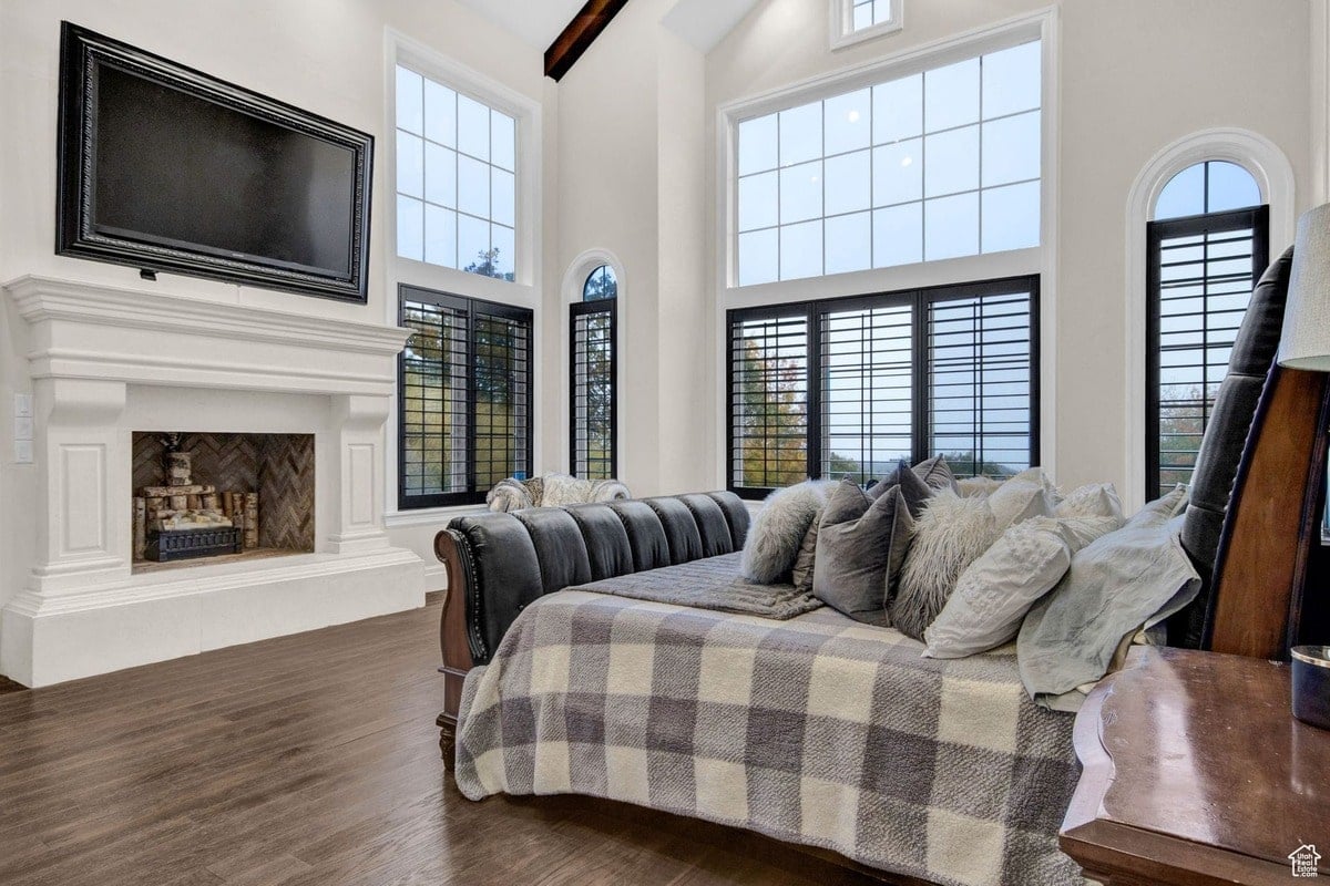 This master bedroom's design is highlighted in the photograph, emphasizing the dramatic effect of the large windows and high ceilings. The luxurious bed and fireplace with integrated television create a focal point, showcasing a blend of comfort and modern technology.