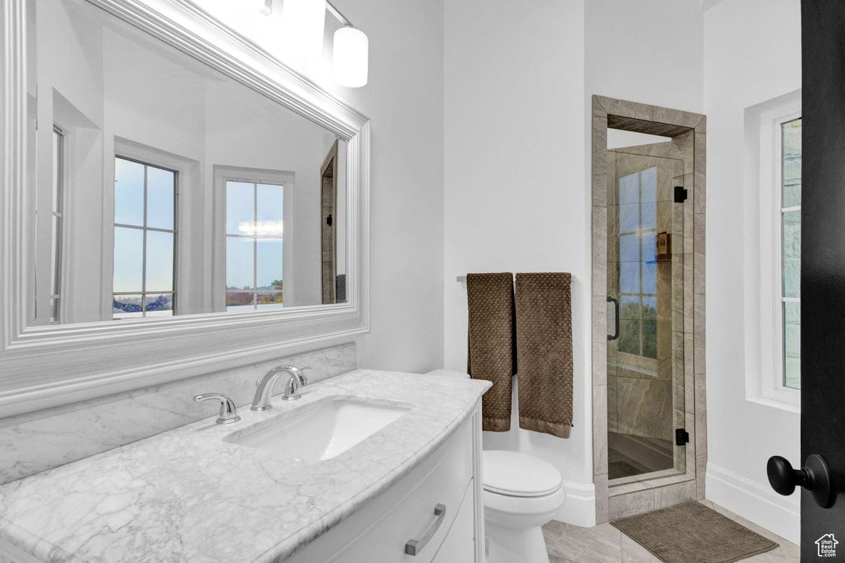 This elegant bathroom boasts a marble-topped vanity with a double sink and an expansive mirror reflecting a view of a window. A glass-enclosed shower is visible in the background, completing the serene and luxurious bathroom design.