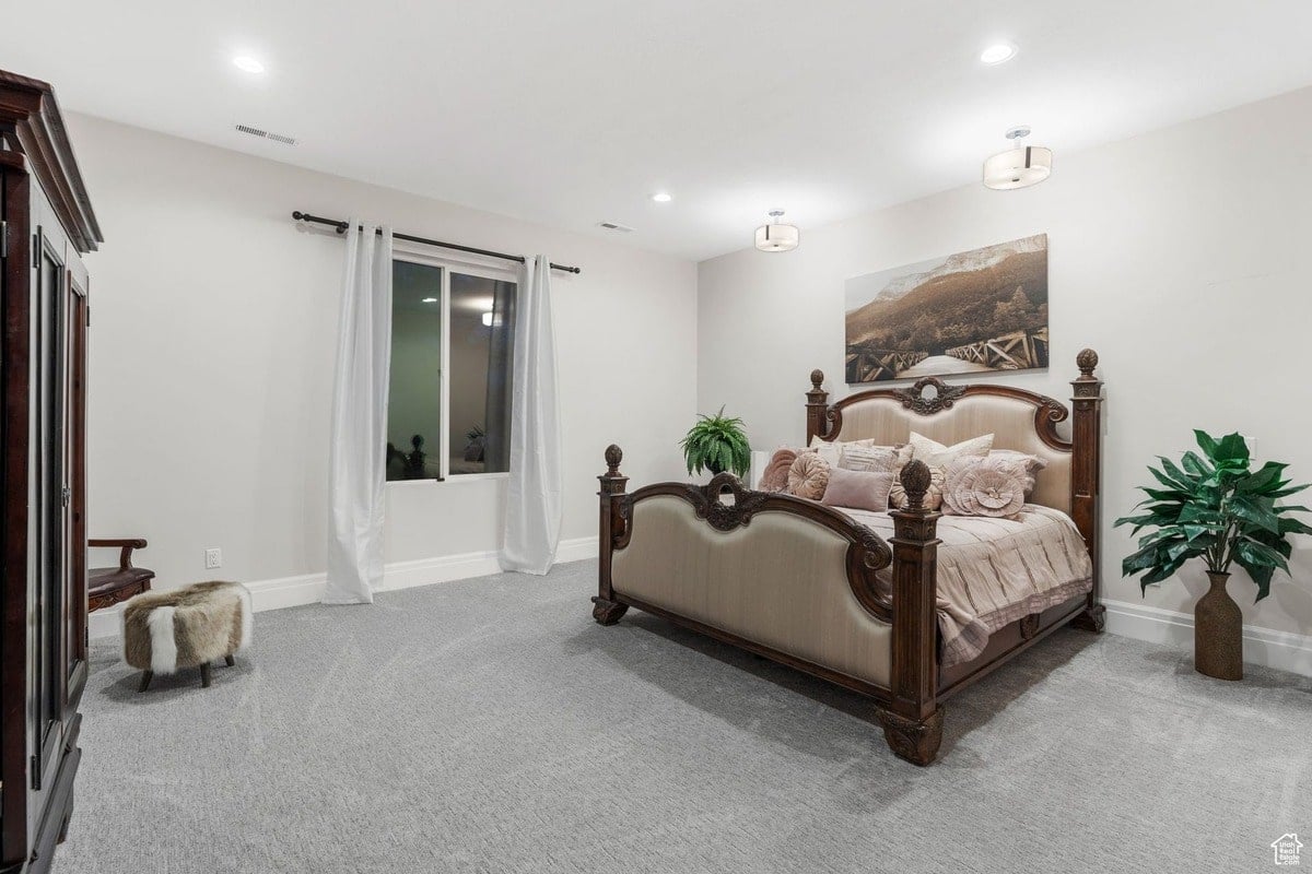 This spacious bedroom features a large, ornate bed with a neutral-toned upholstered headboard, creating a sophisticated and calming atmosphere. The gray carpeting and large window with white curtains enhance the room's airy and restful ambiance.