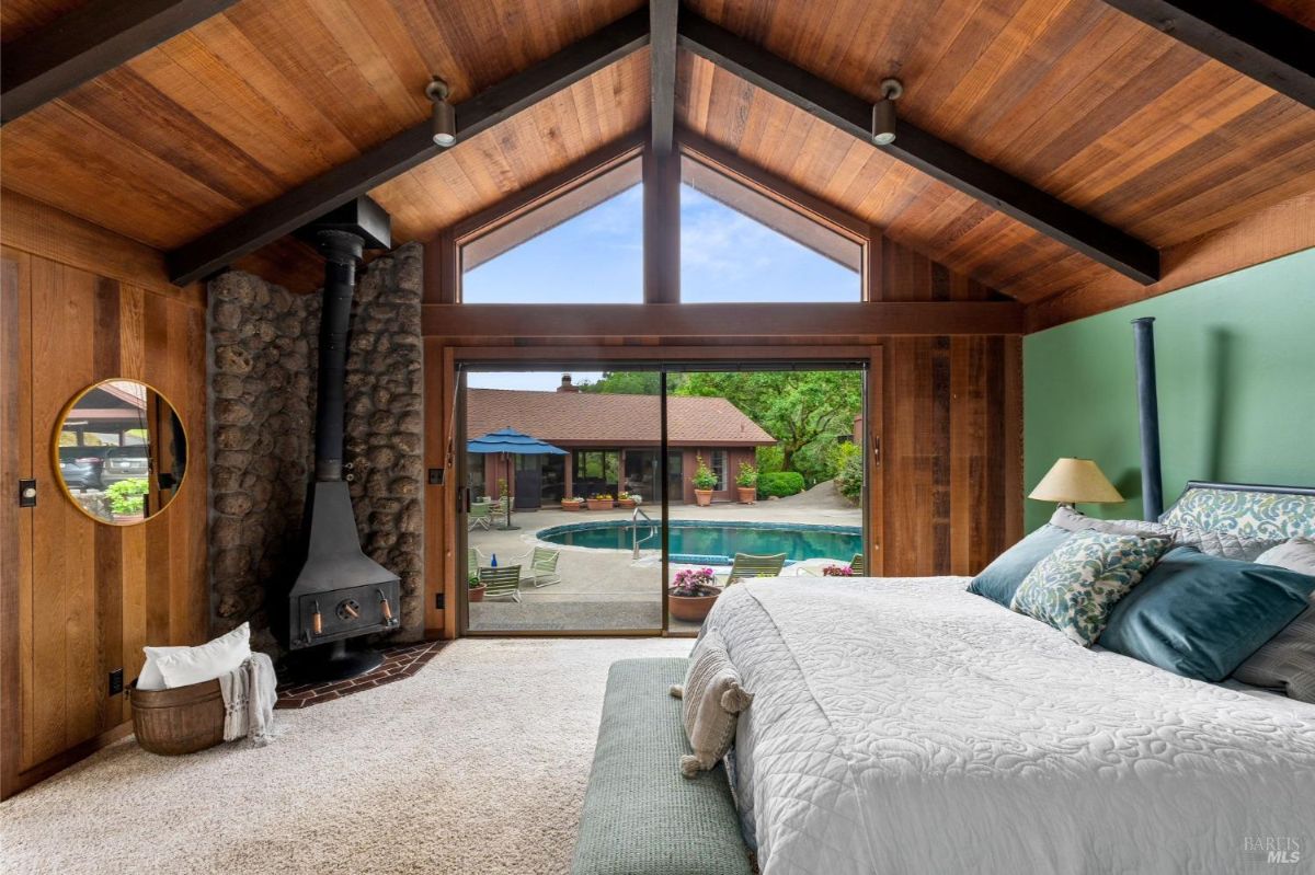 Rustic bedroom features wood-paneled walls, vaulted ceiling, and a view of the pool through large sliding doors.