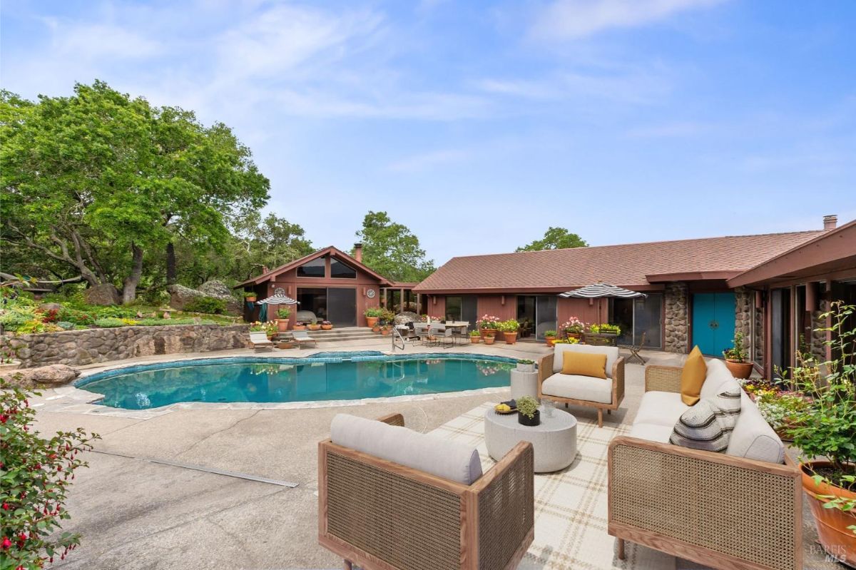 Spacious pool area is surrounded by lush greenery, comfortable seating, and rustic architectural elements.