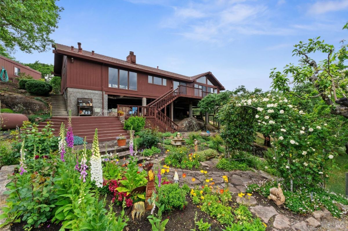Vibrant garden surrounds a red house with a deck and stairs leading to lush greenery.
