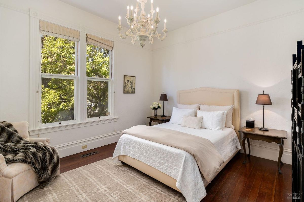 Bright bedroom with a chandelier, light-colored furniture, and large windows overlooking greenery.
