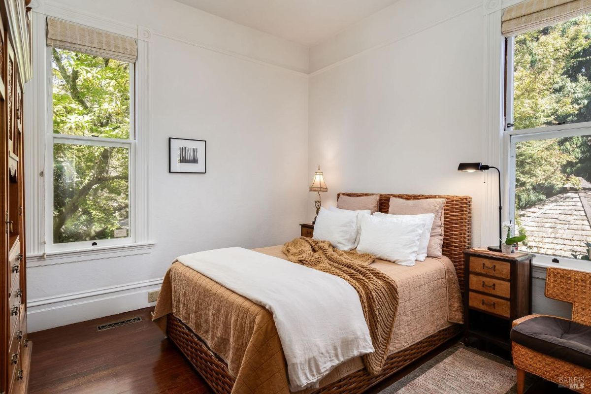 Bright bedroom with wicker bed frame, white bedding, and views of greenery through multiple windows.
