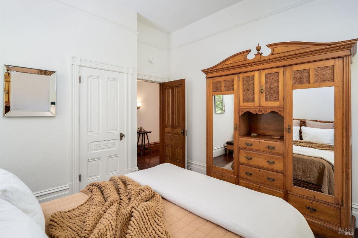 Wooden wardrobe with carved details and mirrored panels in a bedroom with white walls and wooden floors.
