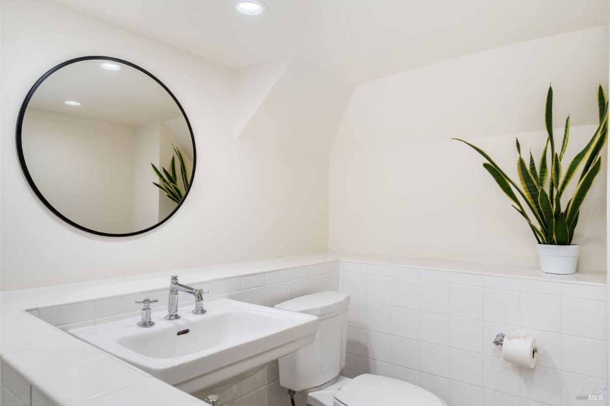 White-tiled bathroom with a wall-mounted sink, circular mirror, and decorative plant.