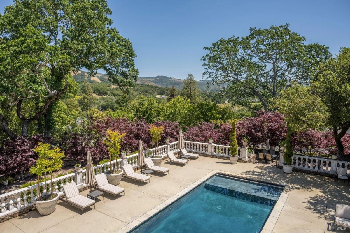 A rectangular swimming pool surrounded by a paved patio area with multiple lounge chairs and umbrellas. White decorative balustrades line the edge of the patio, separating it from the lush greenery and purple foliage beyond. Rolling hills, trees, and a clear blue sky are visible in the background.