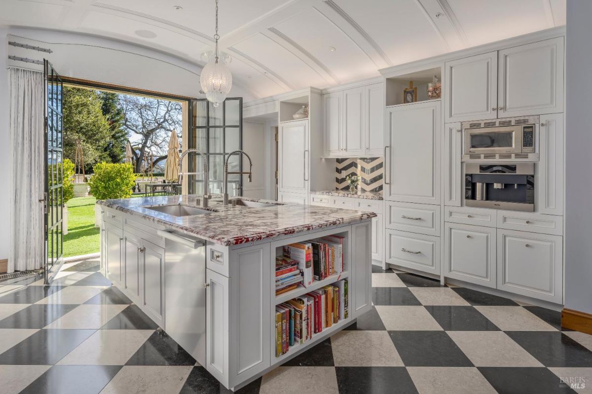 Bright kitchen with a central island featuring marble countertops, double sinks with tall faucets, and shelves holding books. The cabinetry is white with built-in appliances, including a microwave and coffee machine, alongside a decorative chevron-patterned backsplash. Large glass doors open to an outdoor patio and garden area, letting in natural light and providing access to the exterior.