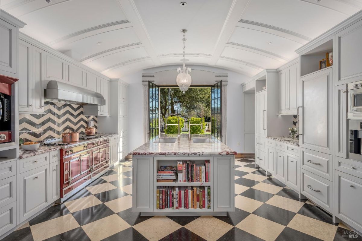 A spacious kitchen with white cabinetry, a central island with a marble countertop, and built-in shelves for books. The backsplash above the stove features a bold chevron pattern, and a large professional-grade range is positioned along the wall. Wide glass doors open to an outdoor area with greenery, allowing natural light into the room.