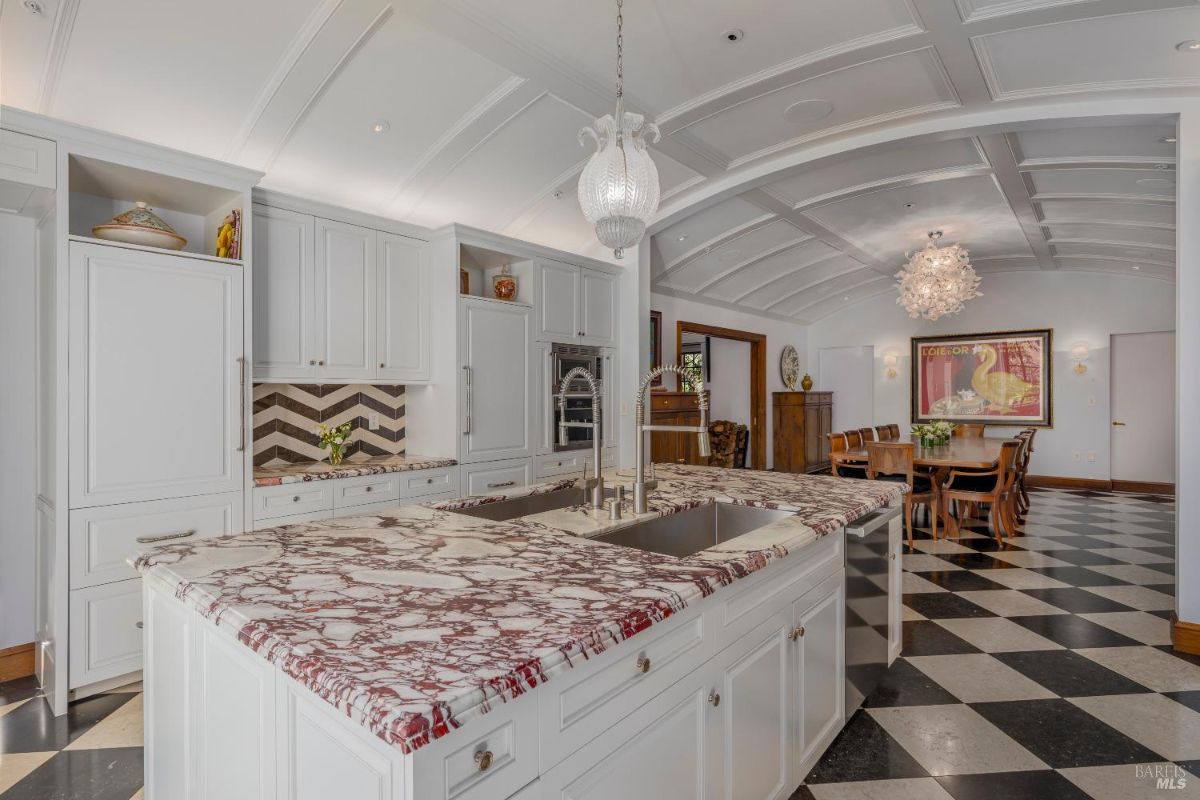 Kitchen with a large marble island that includes dual sinks, two faucets, and ample cabinetry. The space features a white chevron backsplash behind the countertop and built-in cabinets for storage, along with integrated appliances. In the background, an adjoining dining area with a long table and chairs is visible under an arched coffered ceiling, with a chandelier hanging above.