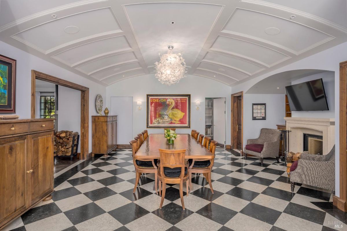 A dining room with a long wooden table surrounded by twelve matching chairs featuring black cushions. The floor has a black-and-white checkered tile pattern, and the ceiling includes a coffered design with a central chandelier. At the far end, there is a fireplace with a mounted TV above it, flanked by two armchairs, while wooden cabinets line the walls for additional storage.