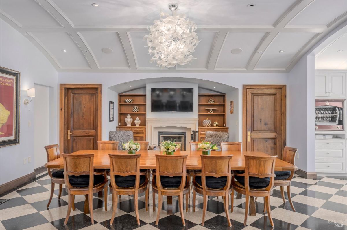 Formal dining room with a large rectangular wooden table surrounded by ten matching wooden chairs with black cushions. The room features a checkered black and white tile floor, a coffered ceiling, and a modern chandelier hanging in the center. At the far end, there is a fireplace below a mounted flat-screen television, flanked by built-in wooden shelves displaying decorative items.
