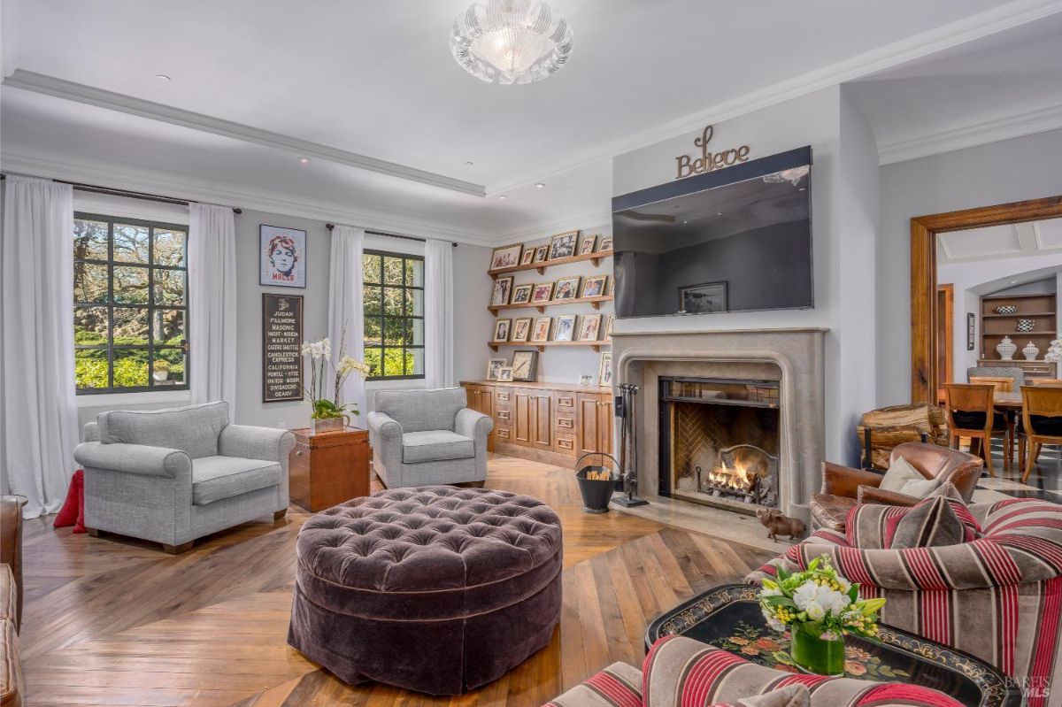 Living room with a lit fireplace, topped by a mounted television and a decorative word sign above it. The space features two gray armchairs, a large round upholstered ottoman, and a striped sofa with floral decorations on the nearby table. Built-in shelves on the back wall display framed photographs, while two large windows with white curtains bring in natural light.