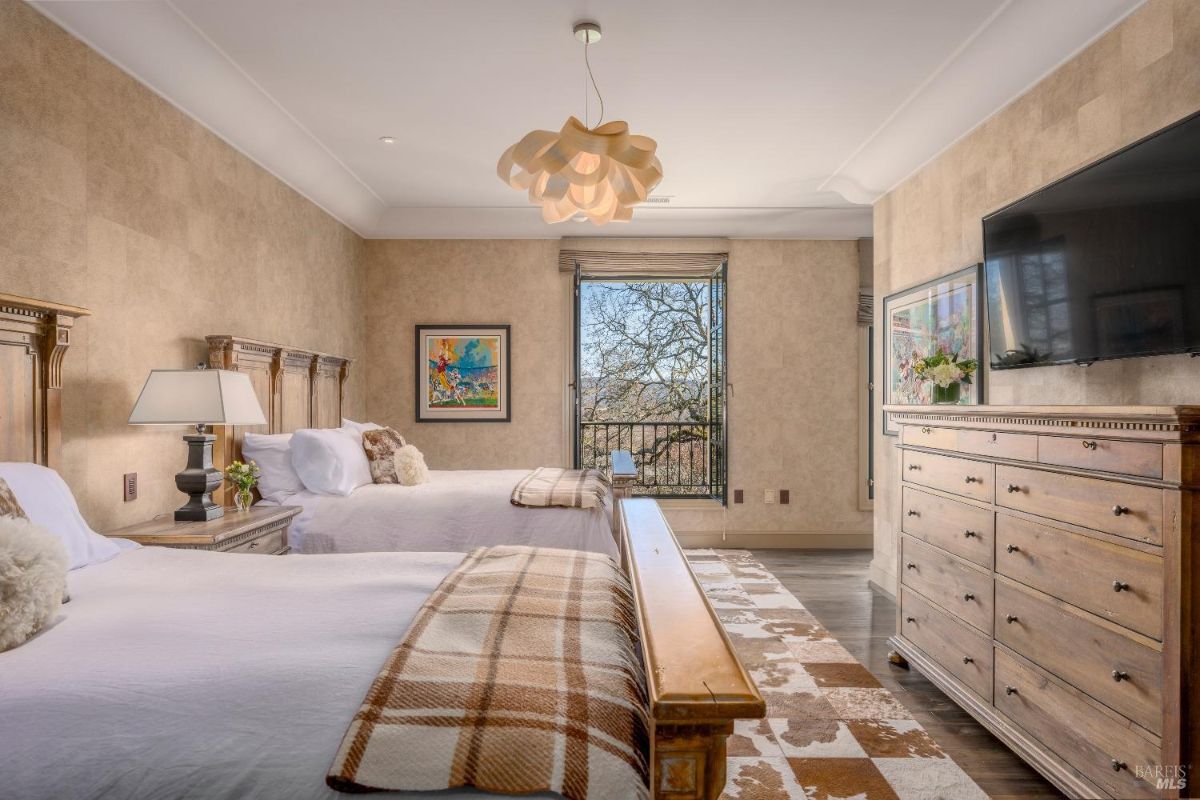 A bedroom featuring two large wooden-framed beds with white bedding and plaid blankets. A wooden dresser with multiple drawers is placed against the wall, beneath a mounted flat-screen TV. The room includes an open window with a view of trees, beige wallpaper, and a modern pendant light hanging from the ceiling.