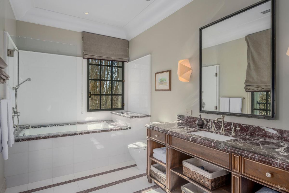 A bathroom with a built-in bathtub featuring marble accents and a glass shower partition. A dark wood vanity with a marble countertop, storage shelves, and a rectangular sink is visible along with a large mirror above it. A window with a beige shade allows natural light to enter, and the space includes a wall-mounted light fixture and framed artwork.