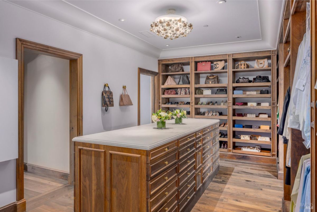Walk-in closet with a central wooden island topped with a white surface and multiple drawers. Built-in wooden shelves line the walls, holding bags, shoes, and clothing neatly arranged. A decorative light fixture hangs from the ceiling, illuminating the organized space.