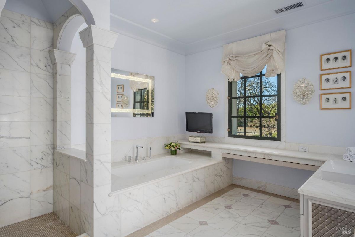 Bathroom featuring a marble soaking tub surrounded by white marble tiles. There is a built-in vanity counter beneath a window, which is dressed with a decorative curtain. A small wall-mounted television and a mirror with built-in lighting are visible on the walls.
