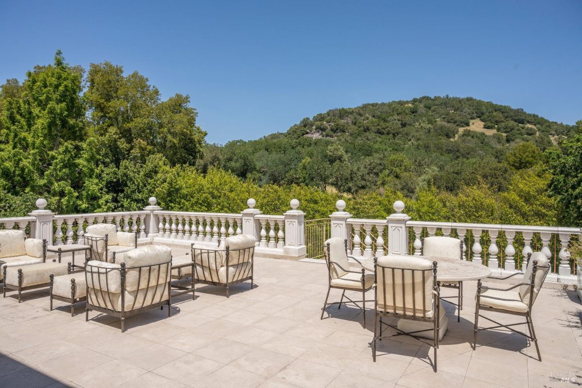 Outdoor terrace with multiple seating arrangements, including cushioned chairs, tables, and lounge furniture. The terrace features a decorative white balustrade surrounding the space. The background consists of lush green trees and a rolling hillside under a clear blue sky.