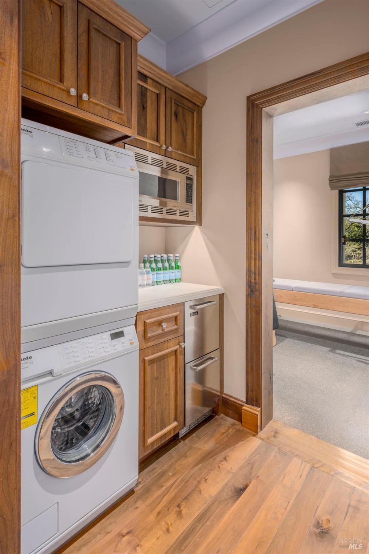 Laundry area with a stacked washer and dryer unit on the left side. Adjacent to the appliances, there is a countertop with wooden cabinetry above and below, as well as a small microwave installed. A narrow stainless steel refrigerator or freezer is positioned under the counter, and a doorway leads into another room with a window and a bench.