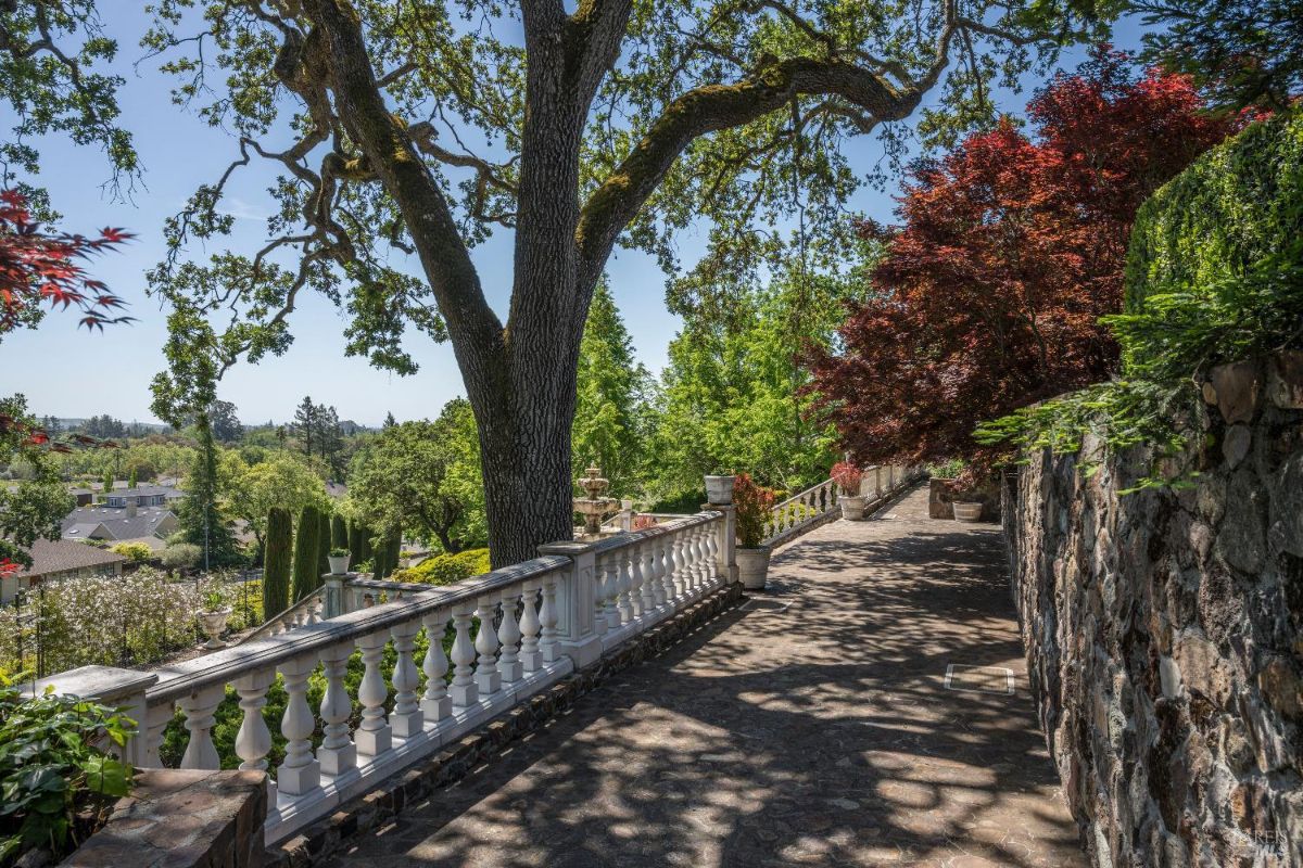 Stone-paved pathway lined with a white balustrade on one side and a rustic stone wall on the other. Large, mature trees with sprawling branches provide shade, while red and green foliage adds vibrant color to the scene. In the background, there are distant treetops, a fountain, and partial views of rooftops, suggesting a garden or estate setting.