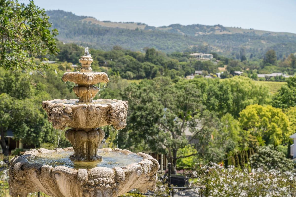 Multi-tiered stone fountain with water flowing gently from its tiers. Surrounding the fountain is a lush landscape with trees, shrubs, and a distant view of rolling hills and greenery. The fountain sits prominently in the foreground, contrasting with the expansive natural backdrop.