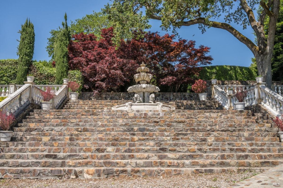 Multi-tiered stone staircase leading up to a three-tiered fountain situated in the center. On either side of the staircase are white stone railings adorned with potted plants. Behind the fountain, there are red and green trees, with additional landscaping and a large tree to the right providing shade.