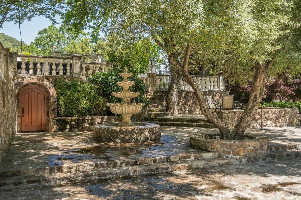 A multi-tiered stone fountain in a courtyard with surrounding stone surfaces and steps. A large tree with a thick trunk and sprawling branches provides shade, and the courtyard is bordered by stone walls and balustrades. A wooden arched door is set into the stone wall, and lush greenery surrounds the space.